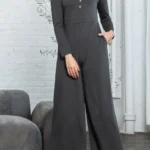 Trinity Button Long Sleeve Wide Leg Jumpsuit - A chic and versatile jumpsuit featuring a button-down front, long sleeves, and wide-leg pants, perfect for creating a stylish and effortlessly elegant look for any occasion. Background of small gray sofa, a indoor tree and white brick wall.