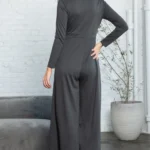 Trinity Button Long Sleeve Wide Leg Jumpsuit - A chic and versatile jumpsuit featuring a button-down front, long sleeves, and wide-leg pants, perfect for creating a stylish and effortlessly elegant look for any occasion .Lady displaying back of jumpsuit. Background of small gray sofa, a indoor tree and white brick wall.
