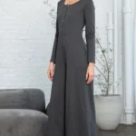Trinity Button Long Sleeve Wide Leg Jumpsuit - A chic and versatile jumpsuit featuring a button-down front, long sleeves, and wide-leg pants, perfect for creating a stylish and effortlessly elegant look for any occasion. Background of small gray sofa, a indoor tree and white brick wall.