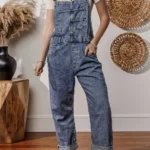 Piper Denim Straight Leg Jumpsuit with Pockets - A casual yet chic denim jumpsuit featuring a straight leg cut, practical pockets, and a timeless design, ideal for effortless everyday style .Woman standing in front of white wall with oval straw wall hangings, and table with pampas grass in black vase,