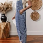 Piper Denim Straight Leg Jumpsuit with Pockets - A casual yet chic denim jumpsuit featuring a straight leg cut, practical pockets, and a timeless design, ideal for effortless everyday style .Woman standing in front of white wall with oval straw wall hangings, and table with pampas grass in black vase.