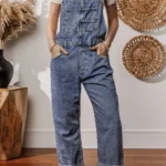 Piper Denim Straight Leg Jumpsuit with Pockets - A casual yet chic denim jumpsuit featuring a straight leg cut, practical pockets, and a timeless design, ideal for effortless everyday style .Woman standing in front of white wall with oval straw wall hangings, and table with vase.