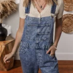 Piper Denim Straight Leg Jumpsuit with Pockets - A casual yet chic denim jumpsuit featuring a straight leg cut, practical pockets, and a timeless design, ideal for effortless everyday style .Woman standing in front of white wall with oval straw wall hangings, and table with vase.