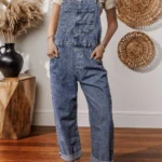 Piper Denim Straight Leg Jumpsuit with Pockets - A casual yet chic denim jumpsuit featuring a straight leg cut, practical pockets, and a timeless design, ideal for effortless everyday style .Woman standing in front of white wall with oval straw wall hangings, and table with pampas grass in black vase.
