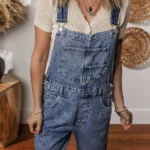 Piper Denim Straight Leg Jumpsuit with Pockets - A casual yet chic denim jumpsuit featuring a straight leg cut, practical pockets, and a timeless design, ideal for effortless everyday style .Woman standing in front of white wall with oval straw wall hangings, and table with vase.