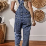 Piper Denim Straight Leg Jumpsuit with Pockets - A casual yet chic denim jumpsuit featuring a straight leg cut, practical pockets, and a timeless design, ideal for effortless everyday style .Woman standing with back turned in front of white wall with oval straw wall hangings, and table with pampas grass in black vase,