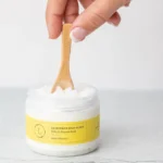 Extended hand holding small wooden spoon, in the process of scooping small amount of Grapefruit Body Scrub from containter. Displayed on white table with white background.