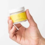 Extended hand holding container of Clay Facial Mask in front of white background.