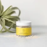 Jar of Clay Facial Mask displayed on white table with clay mask sprinkled around jar . A potted plant and white wall background.